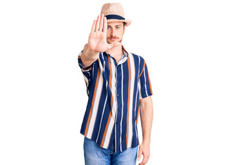 Young handsome caucasian man wearing summer hat doing stop sing with palm of the hand. warning expression with negative and serious gesture on the face.