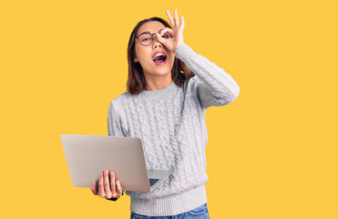 Young beautiful chinese girl wearing glasses holding laptop smiling happy doing ok sign with hand on eye looking through fingers