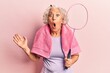 Senior grey-haired woman holding badminton racket wearing towel scared and amazed with open mouth for surprise, disbelief face