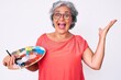 © Krakenimages.com - Senior hispanic grey- haired woman holding paintbrush and palette celebrating victory with happy smile and winner expression with raised hands