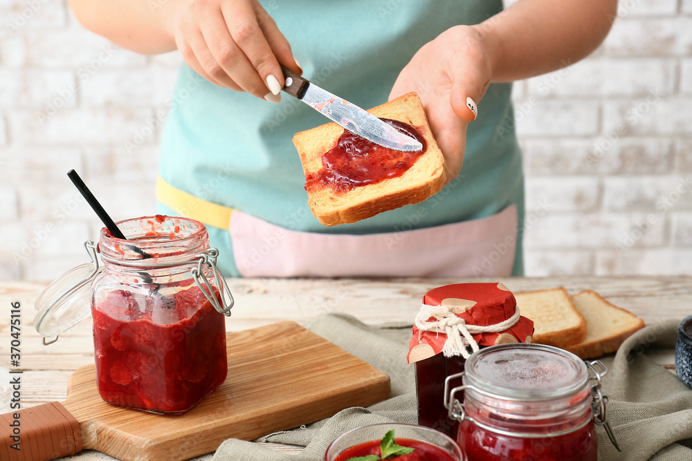 Woman making toasts with sweet strawberry jam in kitchen - obrazy, fototapety, plakaty 