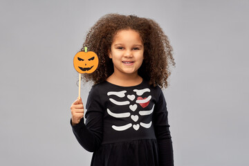 Wall Mural - halloween, holiday and childhood concept - smiling african american girl in black costume dress with skeleton bones and jack-o-lantern party accessory over grey background