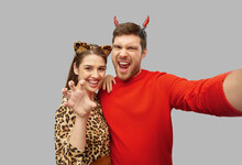 Holiday And People Concept - Happy Couple In Halloween Costumes Of Devil And Leopard Taking Selfie Over Grey Background