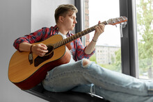 Leisure, Music And People Concept - Young Man Or Musician Playing Guitar Sitting On Windowsill