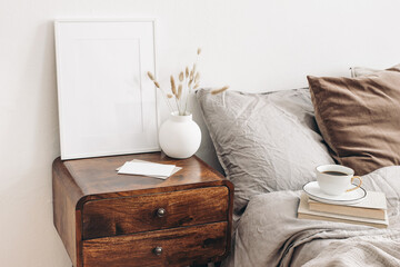Wall Mural - Portrait white frame mockup on retro wooden bedside table. Modern white ceramic vase, dry Lagurus ovatus grass. Cup of coffee and books in bed. Beige linen pillows in bedroom. Scandinavian interior.