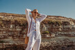 © Victoria Fox - Outdoor summer fashion portrait of elegant woman wearing white linen suit, sunglasses, with round wicker bag, posing on rocks near sea. Copy, empty space for text