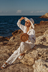 Outdoor summer fashion portrait of elegant woman wearing white linen suit, straw hat, sunglasses, leather sandals, with round wicker bag, posing on rocks near sea