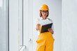 © standret - With notepad in hands. Female worker or engineer in yellow uniform and hard hat standing indoors