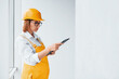 © standret - With notepad in hands. Female worker or engineer in yellow uniform and hard hat standing indoors