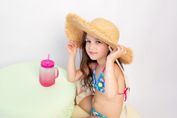 a little girl of 5-6 years old is sitting in a swimsuit with a cocktail on a white isolated background, a place for text