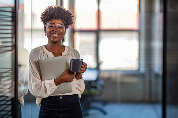 Wall Mural - Pretty young dreamy African-American office worker standing with arms crossed and looking at camera. Career Girl. African American Businesswoman Posing vit laptop and cup od coffee