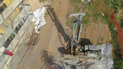 Wall Mural - AERIAL: Flying above a massive machine burrowing a hole in the ground for a heat pump. Heavy machinery digs a hole in the gravel driveway in front of a modern prefabricated house under construction.