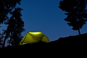 The tent glows in the night. Camping by the lake in a pine forest. Romantic trip.