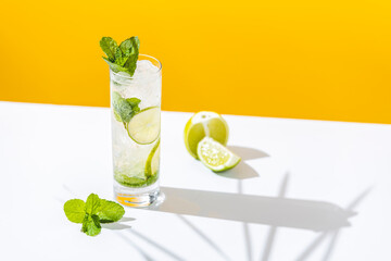 Fresh Mojito cocktail with lime and mint in highball glass with water drops on white and yellow background in bright sun light, Mojito refreshing drinks. Copy space, summer vacation and party concept