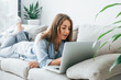 © standret - Beautiful young woman in jeans and blue shirt lying down at sofa with laptop