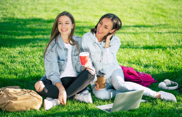 Two happy beautiful young student girl friends in casual denim clothes are relaxing in college park with laptop and smartphone on university background and drinking coffee.