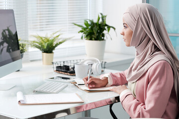 Wall Mural - Busy young Muslim woman in hijab sitting in modern office and making notes while working with data