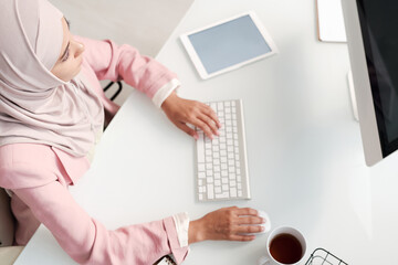 Poster - Above view of busy young Muslim woman in hijab sitting at desk and working on project using computer