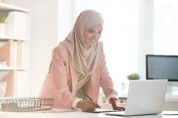 Poster - Positive young Muslim designer in hijab standing at table and editing digital sketch using drawing pad