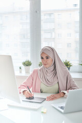 Poster - Busy young Muslim graphic designer in beautiful hijab sitting at desk and using digitizer for work