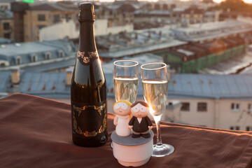 A bottle and two glasses of champagne along with wedding figurines against the backdrop of city rooftops at sunset