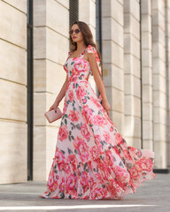 Elegant woman in long pink summer dress with floral print walking at city street. Outdoor full length portrait. Pretty female model