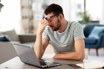 Poster - remote job, technology and people concept - tired young man in glasses with laptop computer working and rubbing his nose bridge at home office