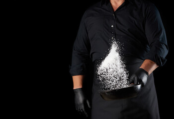 Wall Mural - man in a black uniform holding a round cast iron pan with salt, the chef tosses white salt up