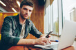 © BullRun - Portrait of male graphic designer looking at camera while installing app on smartphone using 4G internet connection sitting at laptop.Freelancer working at netbook while checking mail on telephone