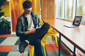Casual dressed young man blogger chatting in social networks on modern smartphone using 4G internet connection.Hipster guy messaging with friends on telephone device resting in coworking space
