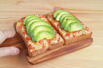 Hand holding a wooden breadboard of grilled cheese toast with tomato and sliced fresh ripe avocado