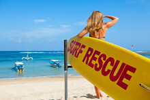Life Saving Yellow Board With Surf Rescue Sign. Young Lifeguard Woman Stand On Duty, Look At Blue Sea. Swimming People Safety. Summer Family Vacation On Ocean Beach. Travel Background.