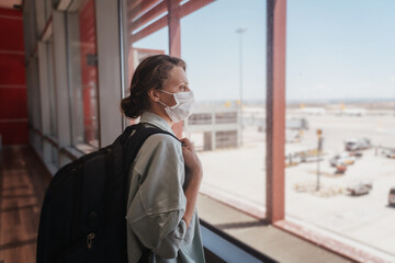 Young woman traveler in a protective medical mask with a backpack looks out the window at the airfield at the airport waiting for boarding a flight