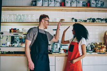 Cheerful Caucasian Barista Giving Five African American Waitress For Great Job During Teamwork In Own Cafeteria.Positive Diverse Male And Female Employees In Aprons Collaborating On Successful Startup