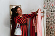 © Antonio Ovejero/ADDICTIVE STOCK - Trendy African American female with braids in striped dress holding high heels and hanger with shirt picking clothes at home