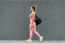 Side View Of Slim Blond Female Athlete In Active Wear And Sneakers With Sports Bag Strolling On Embankment Near Grey Wall In Daylight