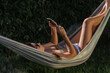 © Fran Rosado/ADDICTIVE STOCK - Side view of cheerful young female in bikini and sunglasses lying in hammock near green plants using smartphone while resting in summer day in garden