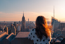 Back View Of Anonymous Female Traveler Standing On Viewpoint Of High Building And Observing Cityscape Of New York City During Sunset