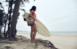 © Luis Gomez/ADDICTIVE STOCK - Side view of young Asian female surfer in summer outfit walking on sandy beach and carrying surfboard against calm blue sea looking away