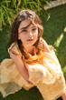 © Philippe Degroote/ADDICTIVE STOCK - From above of adorable calm little girl with long wavy dark hair wearing yellow lace dress sitting on green grass and looking at camera while resting in garden in summer day
