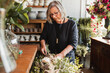 © Raquel Arocena/ADDICTIVE STOCK - Professional adult female florist choosing green decorative plants for composition while working in cozy floristry studio