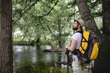 Young Man Doing A Hiking Trail With His Yellow Backpack And Hat On His Head By A Lake With Many Trees And Natural Areas Looking At The Landscape