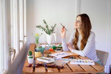 Thoughtful Young Woman Holding Paintbrush While Painting On Table At Home