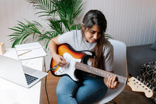 Young Woman Playing Electric Guitar While Sitting On Chair At Home