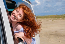 Portrait Of Carefree Redhead Teenage Girl With Head Out Of Car Window At Beach Against Sky