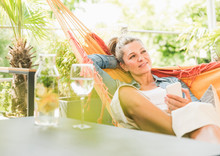 Portrait Of Pensive Mature Woman With Smartphone Relaxing In Hammock On Terrace