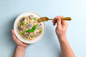 Spaghetti with creamy sauce, champignons and basil leaves on a light blue background. Male hands are holding a plate of Italian pasta and a golden fork. Top view, horizontal orientation.