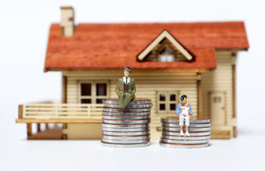 Canvas Print - A miniature man and woman sitting on a pile of coins in front of a miniature wooden house.
