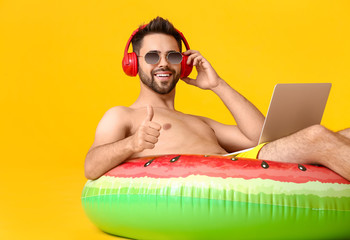 Sticker - Young man with headphones, inflatable ring and laptop showing thumb-up gesture on color background