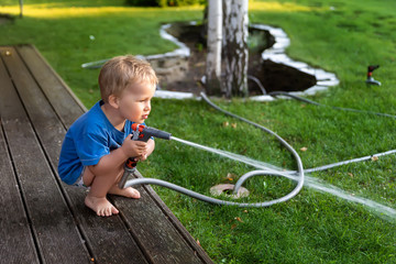 Cute adorable caucasian blond toddler boy enjoy having fun watering garden flower and lawn with hosepipe sprinkler at home backyard at sunny day. Child little helper learn gardening at summer outdoor